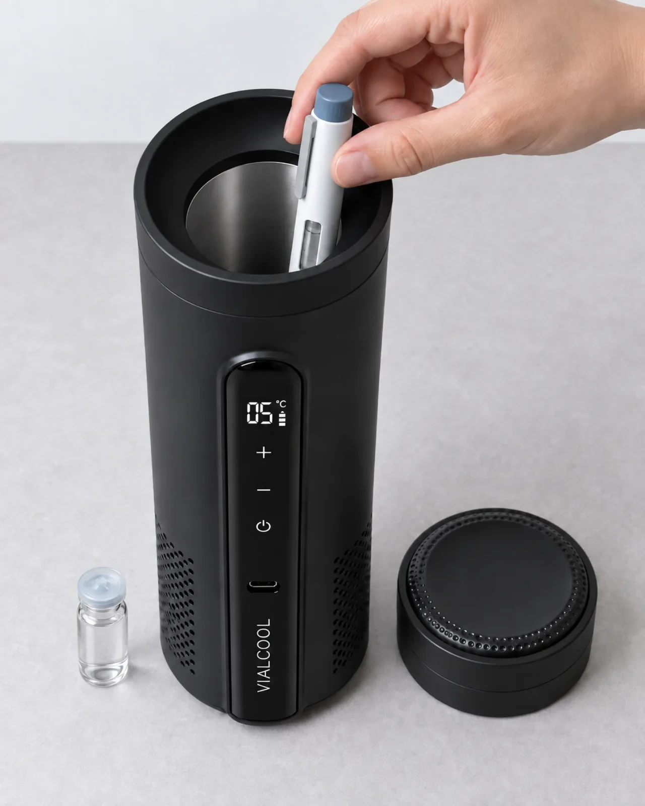 A hand lowering a capped injector pen into the deep silver chamber of the open cooler, a small glass vial standing beside the base, the lid set to one side and the display reading 05°C